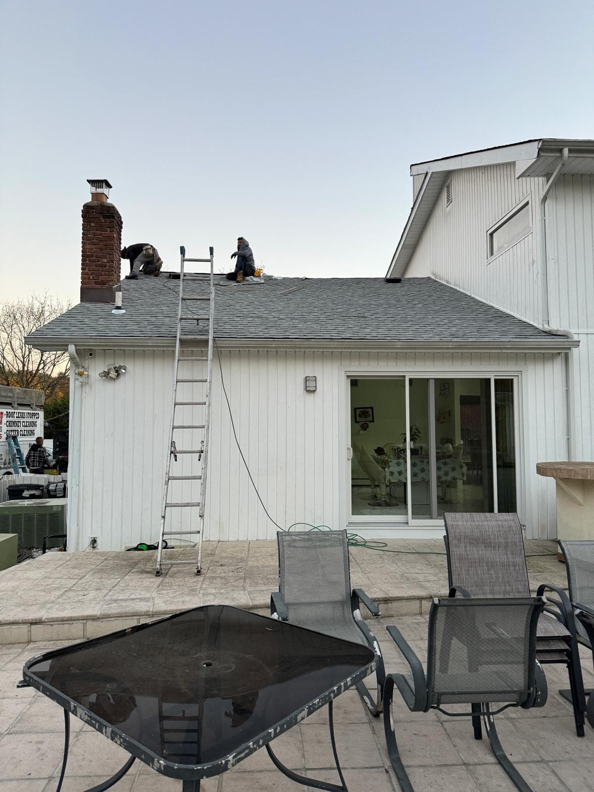 Roofers laying new gray shingles on the same ranch house roof, a ladder against the wall