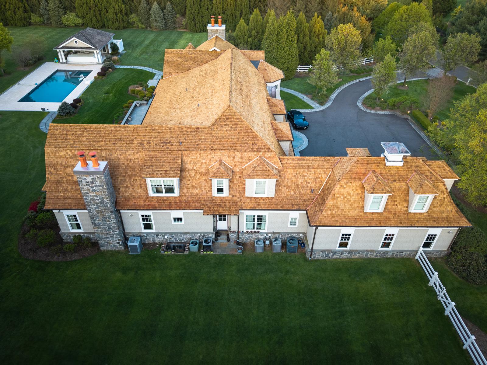 Cedar shake roof with dormers and a stone chimney on a white house beside a pool, seen from above