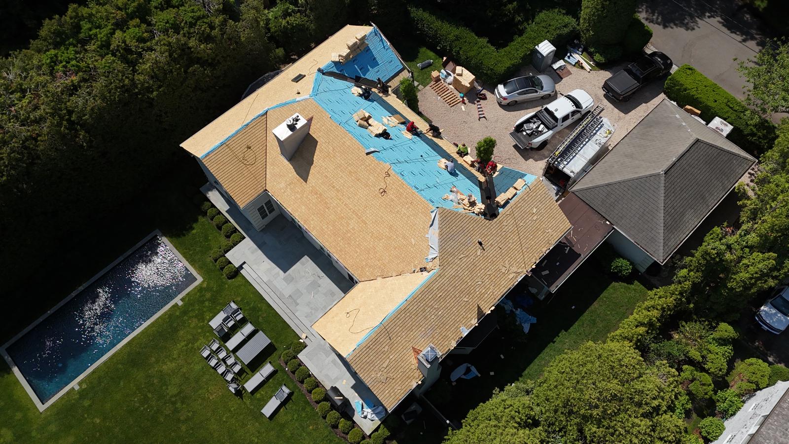 Aerial view of a roof replacement in progress, new blue underlayment going on beside a backyard pool